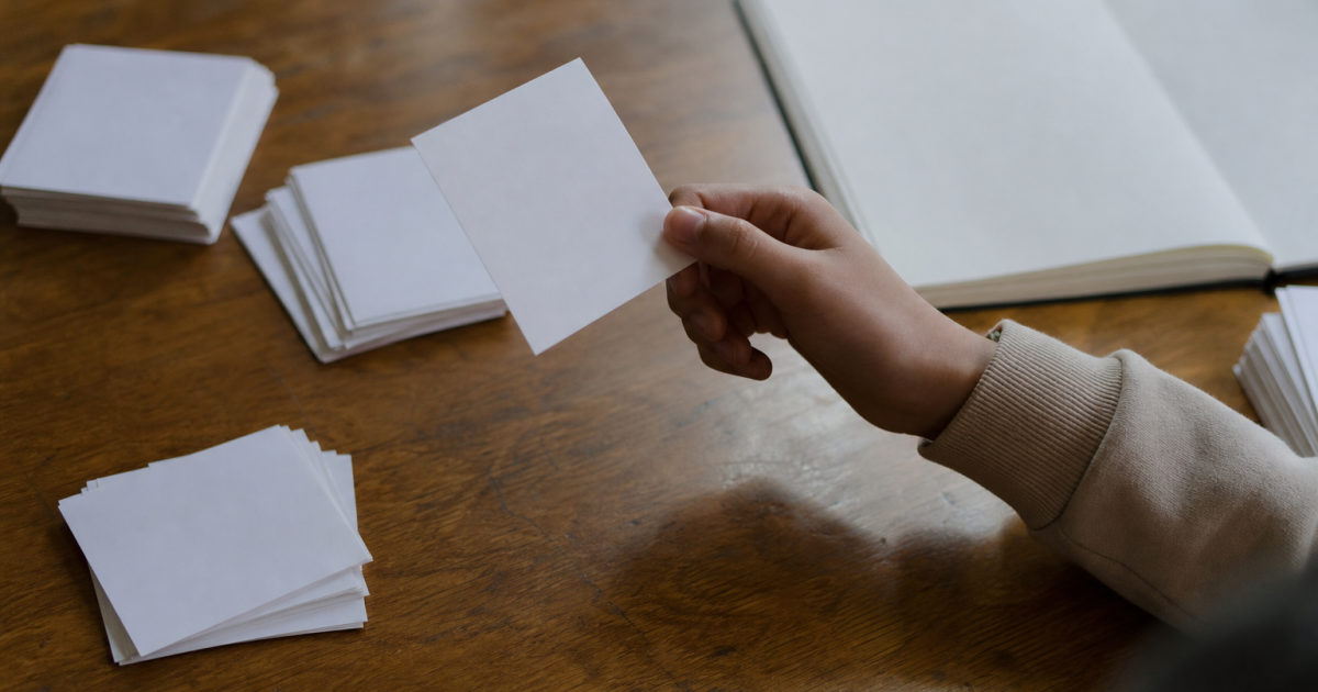 A hand holds a paper flashcard above several card piles beside an open notebook.