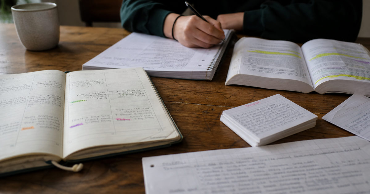 Student seated at a kitchen table with a filled calendar, annotated papers, notes and an open textbook.