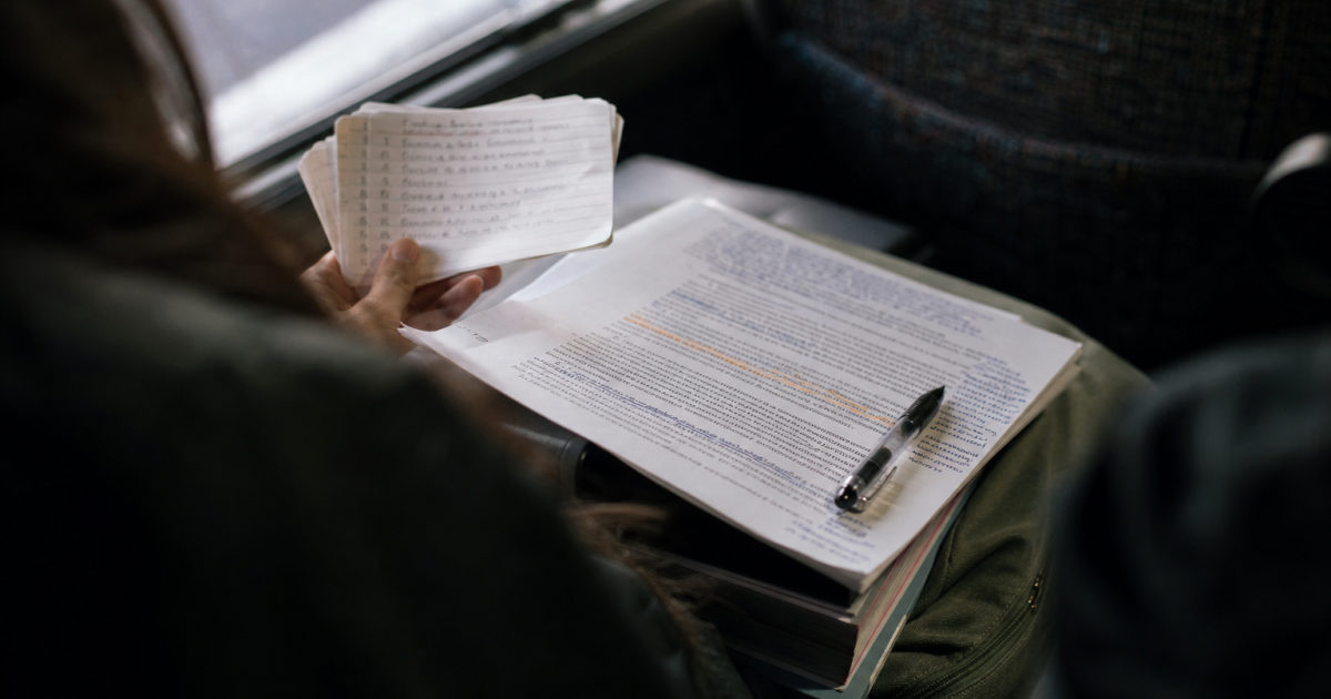 Student on a bus holding handwritten flashcards above an annotated document and a bag of books.