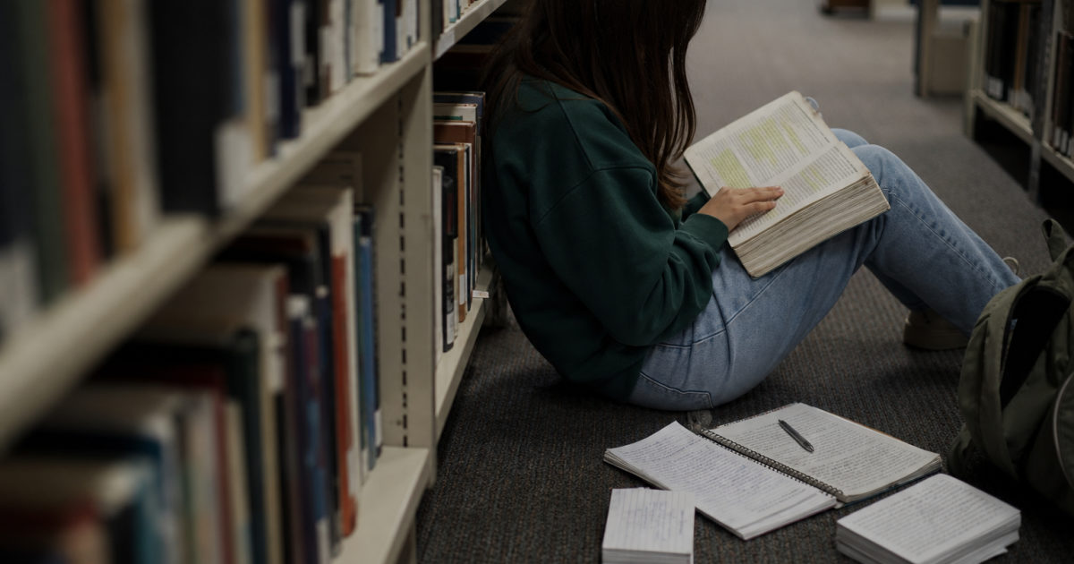 A student sits on the floor in a library aisle with an open highlighted textbook and a handwritten notebook.