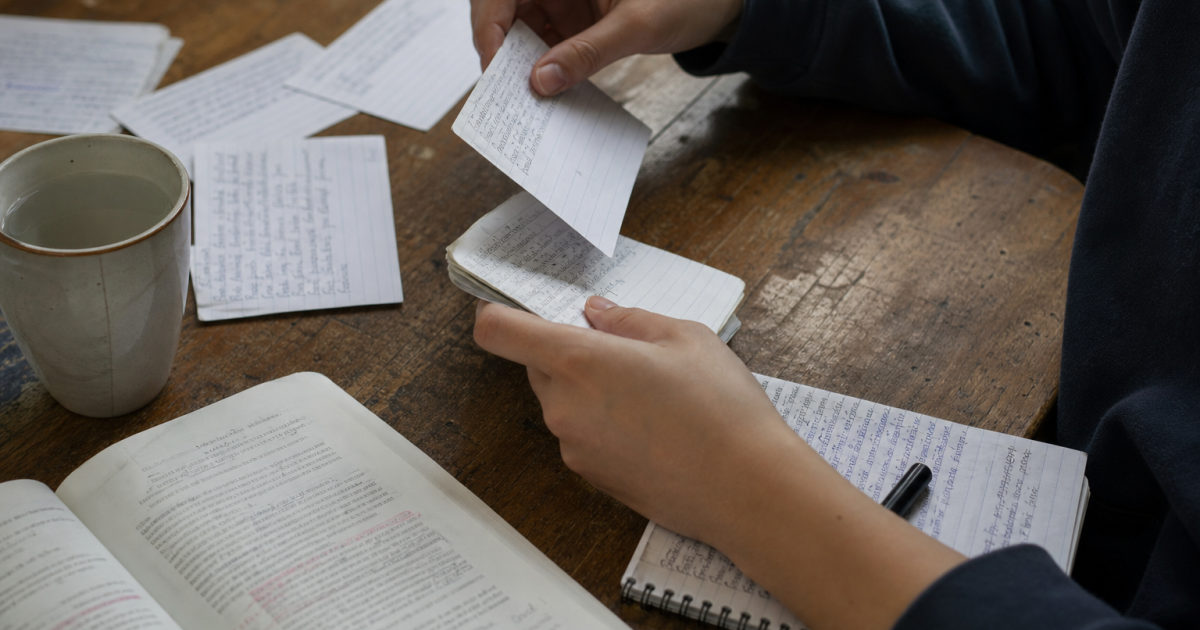 Hands sort written flashcards beside an annotated textbook and a mug on a kitchen table.