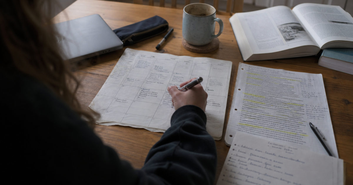 Student marking a weekly calendar beside an annotated assignment brief and open textbooks at a kitchen table.