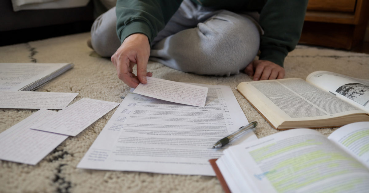 Student organizing handwritten source cards beside an annotated draft and highlighted textbooks on a bedroom floor.