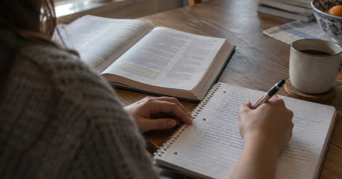 A student's hands write in a partially filled notebook beside an open, highlighted textbook on a kitchen table.
