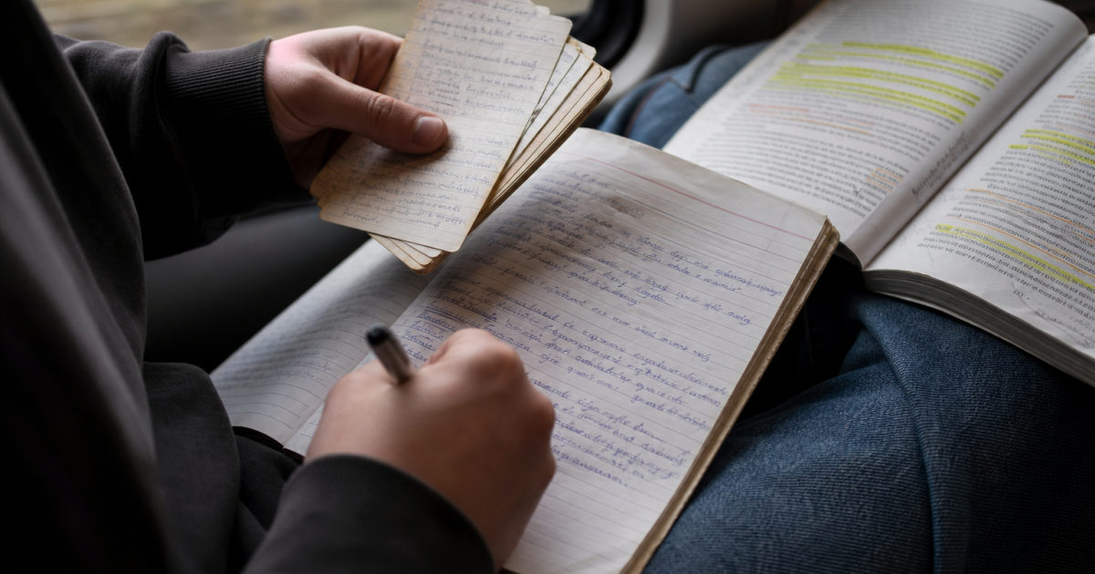 A student on a train writes in a part-filled notebook while holding a stack of handwritten revision cards.