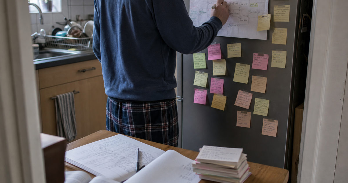 A student stands at a kitchen fridge covered with written sticky notes and updates a calendar filled with revision entries.