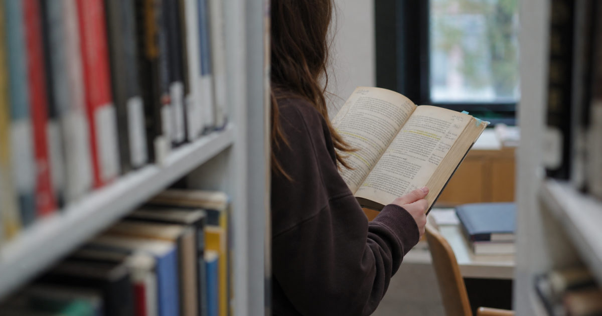 A student stands in a library aisle and previews an annotated textbook held open in both hands.