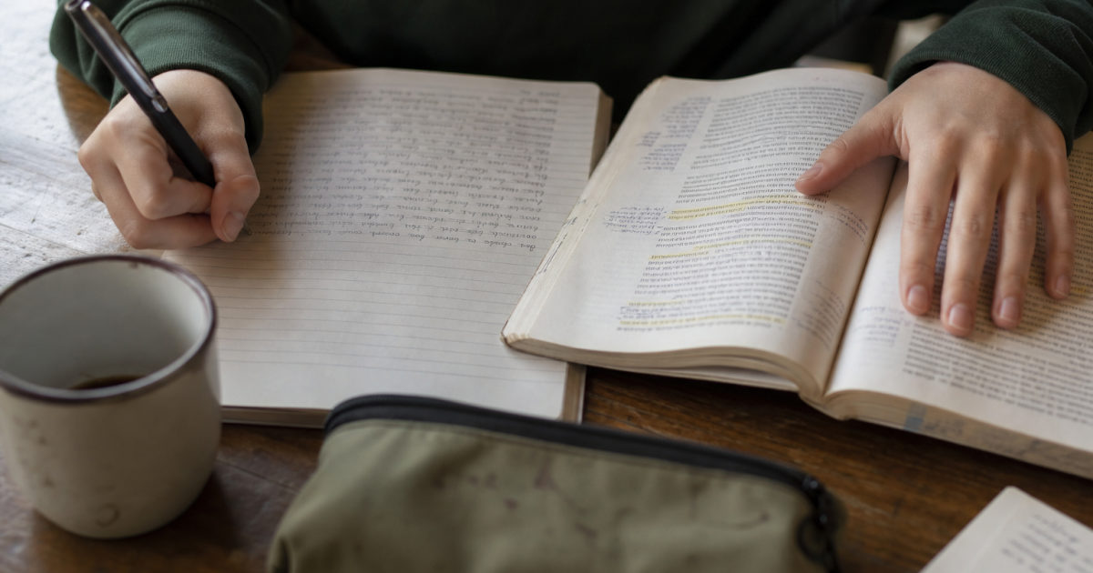 Hands write in a part-filled notebook beside an open textbook with highlighted and annotated passages on a kitchen table.