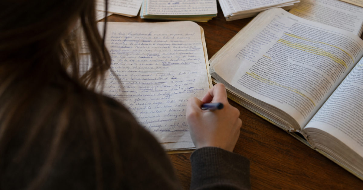A student’s hands mark corrections on a handwritten essay draft beside an open annotated textbook at a library table.
