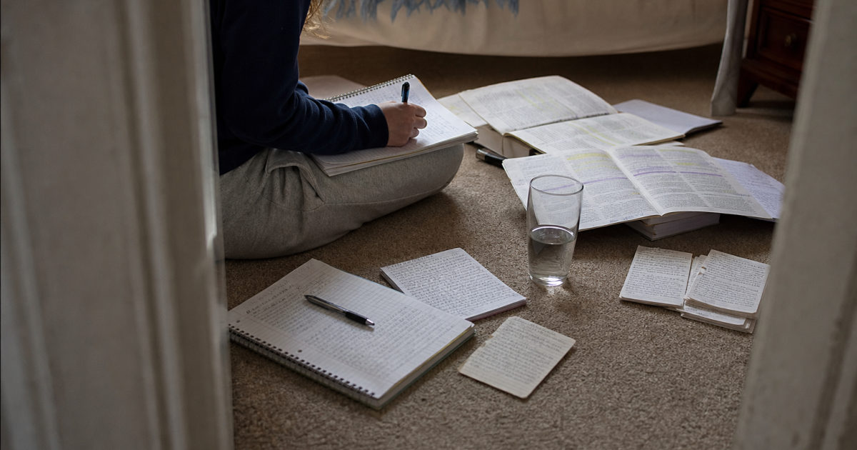 Wide view of a student sitting on a bedroom floor among open annotated textbooks, part-filled notebooks and handwritten revision cards.