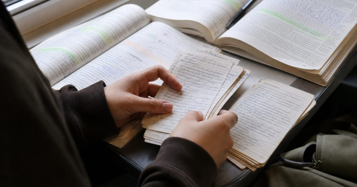 Close view of a student's hands alternating between handwritten cards and two annotated textbooks on a train table.