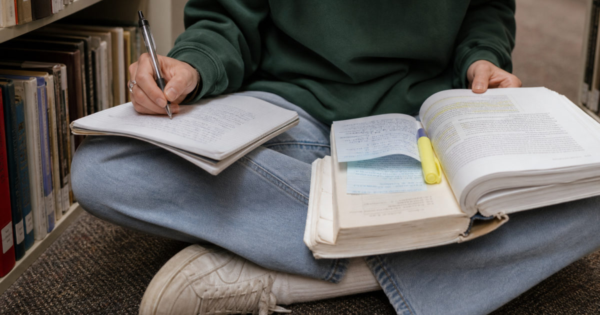 A student sits on a library floor between shelves, comparing a heavily highlighted textbook chapter with part-filled handwritten notes resting on their knees.