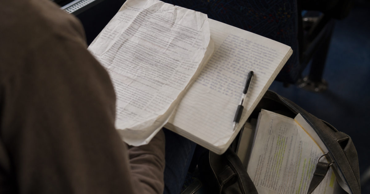 A student seen from behind sits on a bus with an annotated assignment sheet on their lap, a partly filled notebook beside it, and used textbooks tucked into a bag at their feet.