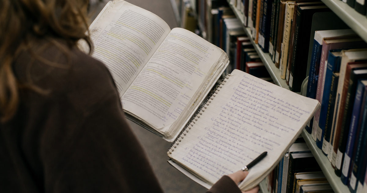 A student in a library aisle holds an annotated textbook beside a handwritten notebook.
