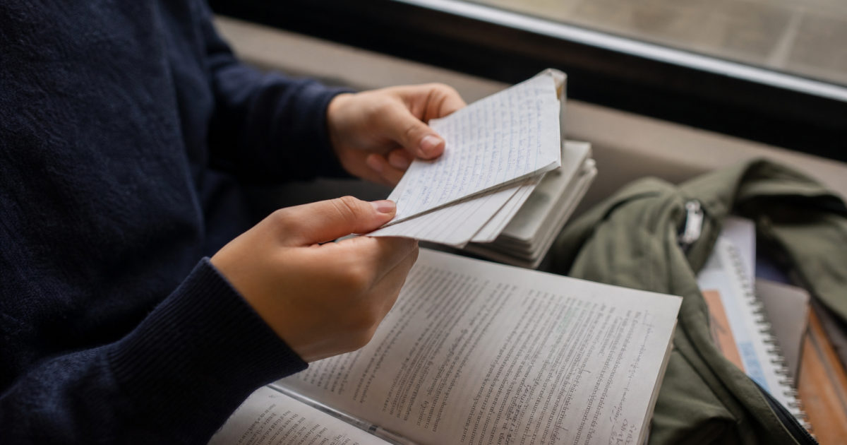 Hands hold handwritten flashcards beside an annotated textbook on a train seat.