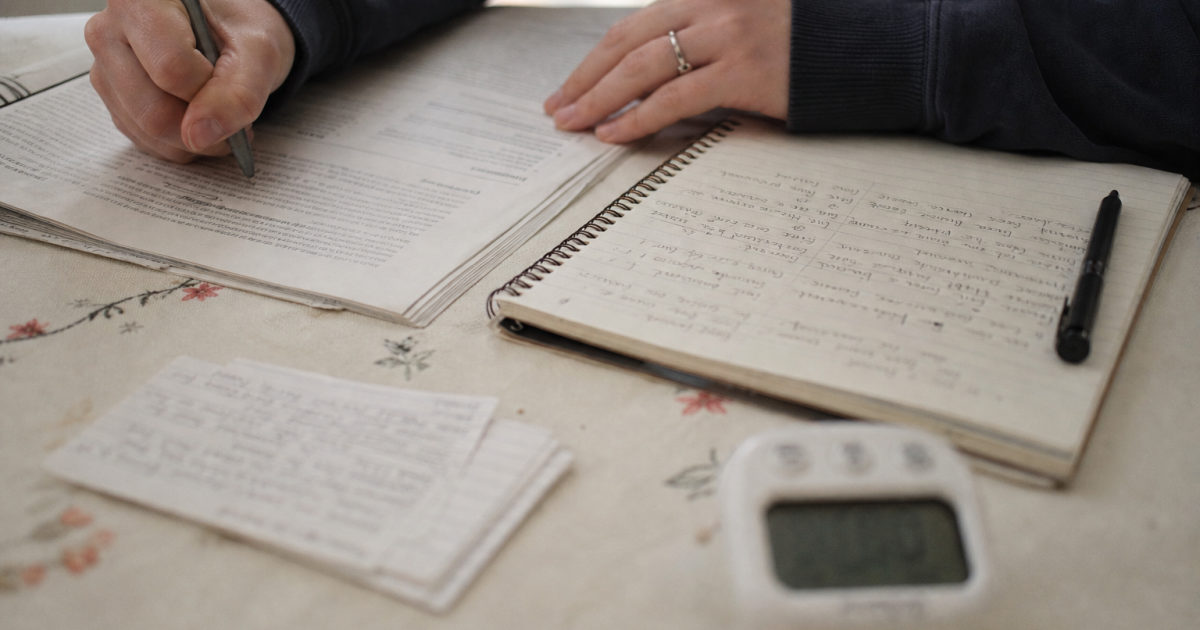 Hands marking a printed assignment brief beside a handwritten notebook and a kitchen timer.
