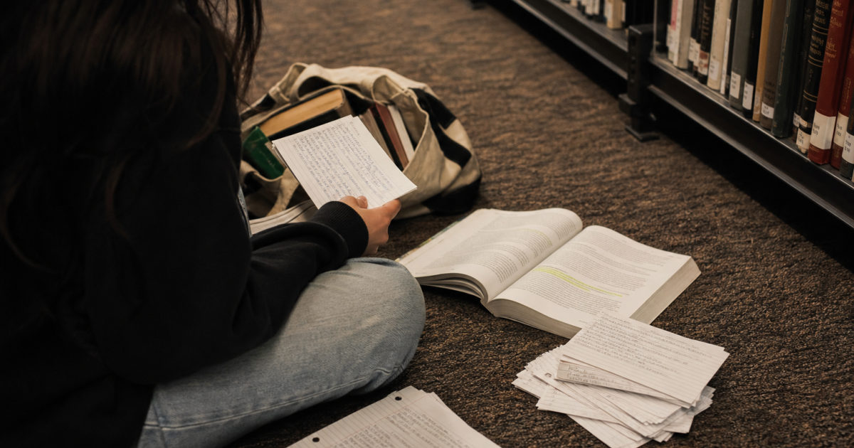 A student sits in a library aisle with an annotated textbook, handwritten cards, printed pages, and a bag of books.