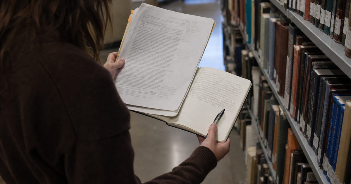 A student in a library aisle holds an annotated article beside a handwritten notebook.