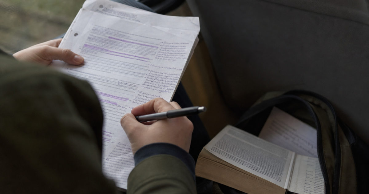 Hands hold an annotated printed essay on a bus beside a pen and a backpack containing a book.