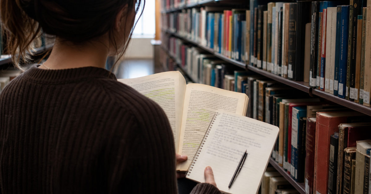 Student in a library aisle holding a highlighted textbook and a notebook filled with handwritten notes.