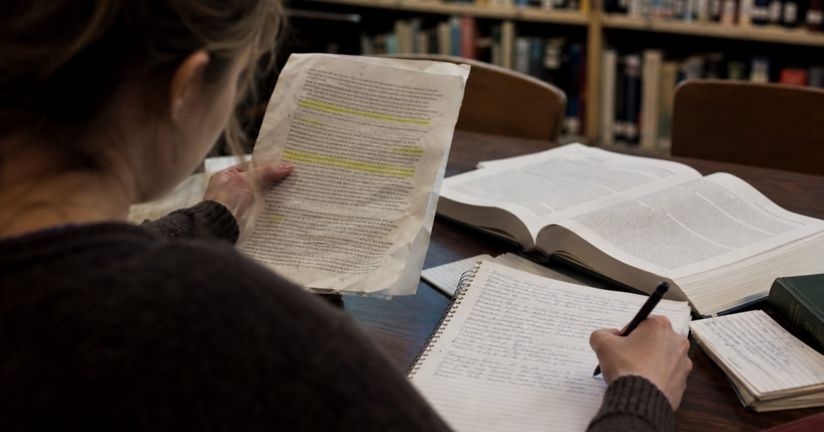 A student at a library table highlights a printed research paper and writes notes in a notebook.