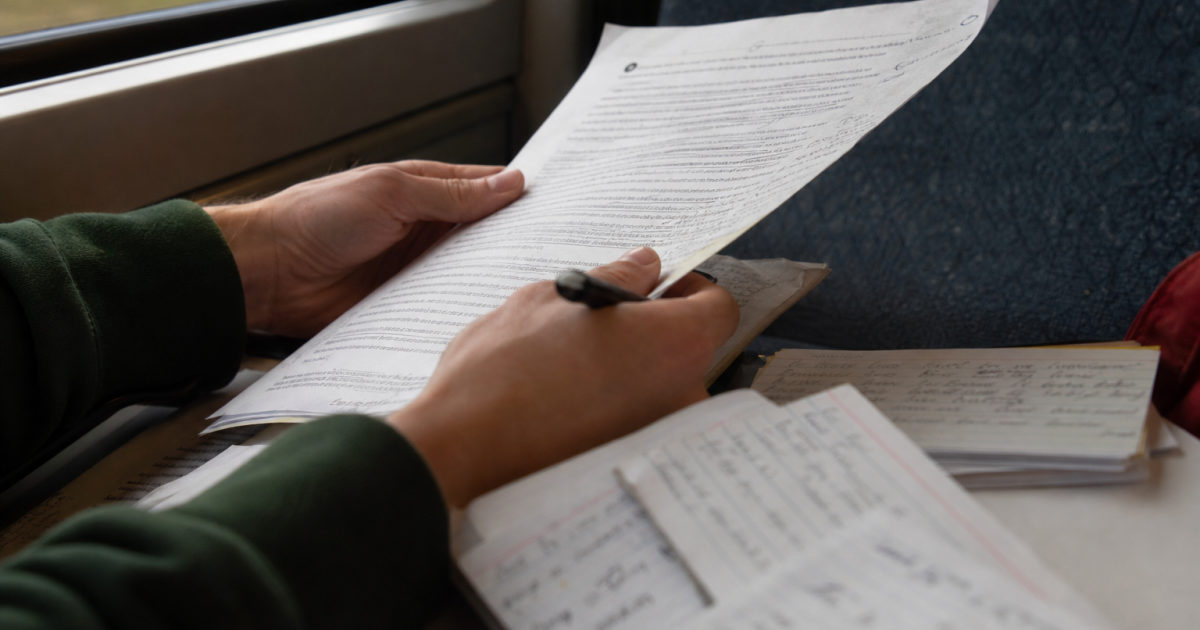 Hands hold an annotated research paper beside handwritten index cards on a train.