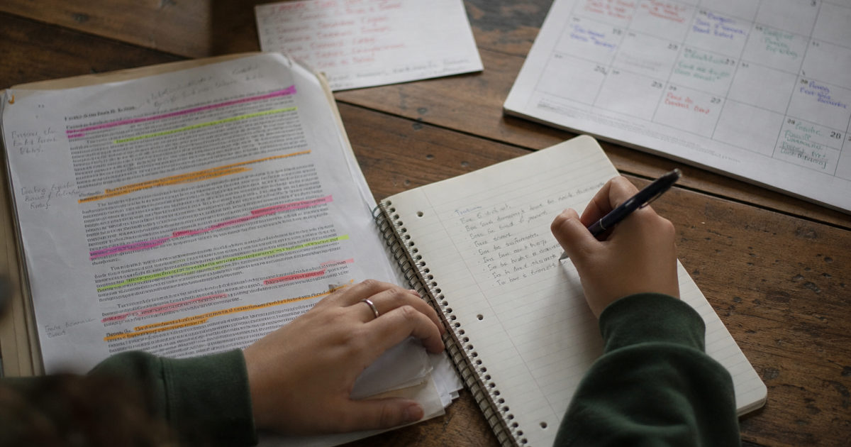 Hands beside a highlighted assignment brief, a partly filled notebook, and a calendar on a kitchen table.