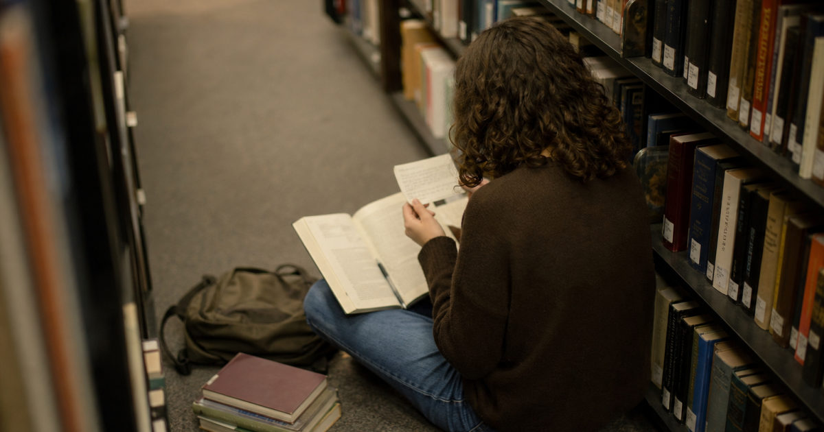 Student viewed from behind sitting in a library aisle with an annotated textbook, handwritten note cards, and a stack of books.