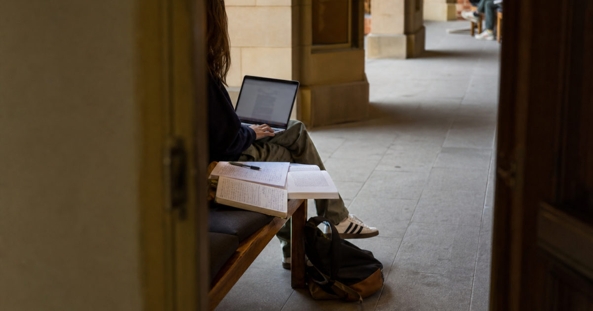 Student sitting on a corridor bench with a laptop, annotated assignment brief, and handwritten notebook.