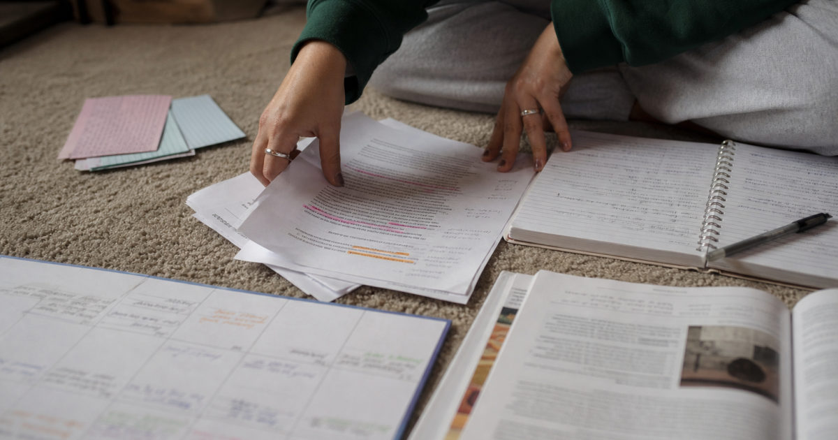 Hands arranging highlighted research pages, an assignment brief, a handwritten notebook, and a filled calendar on a bedroom floor.