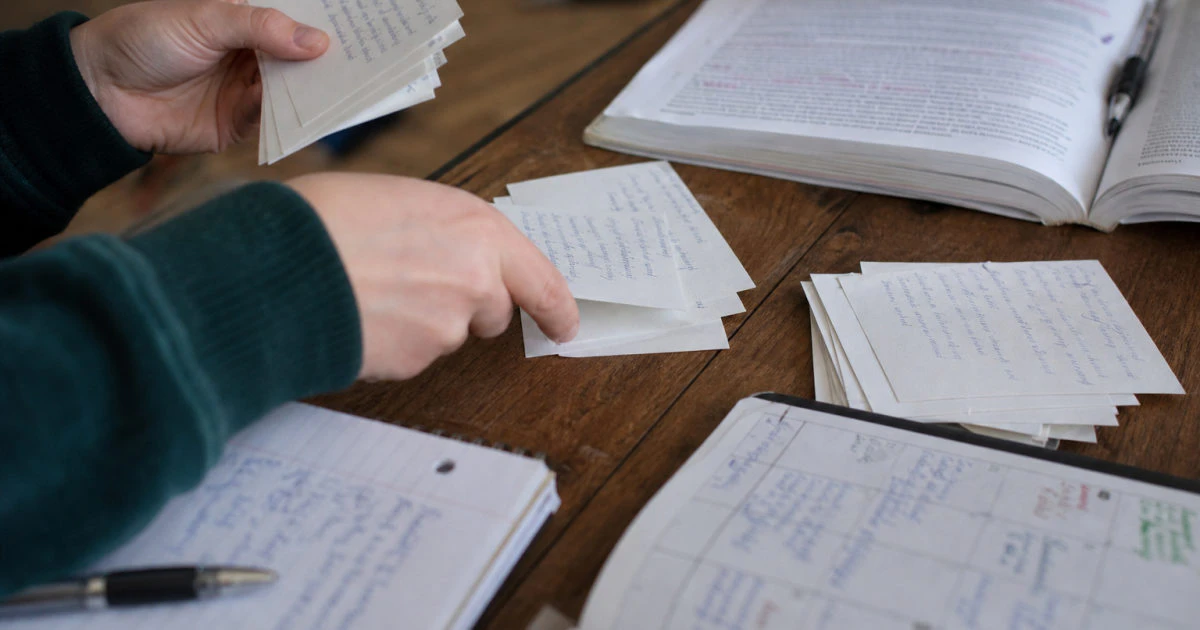 Hands sorting handwritten flashcards beside an annotated textbook and a calendar filled with revision entries.