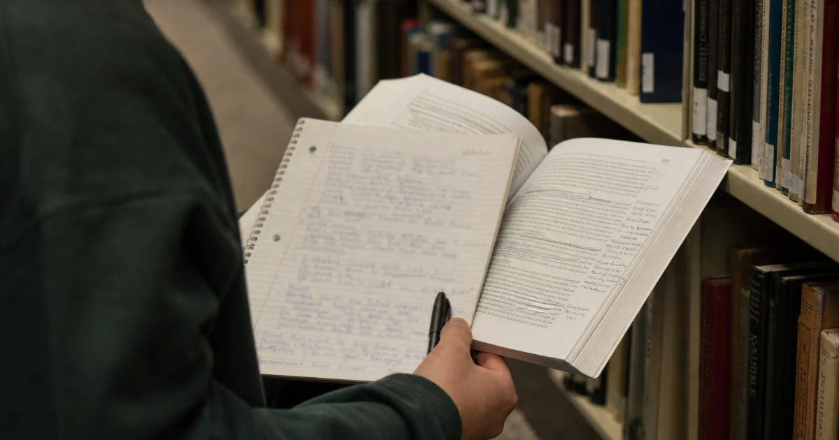 Student in a library aisle holding an annotated textbook and a handwritten topic list.