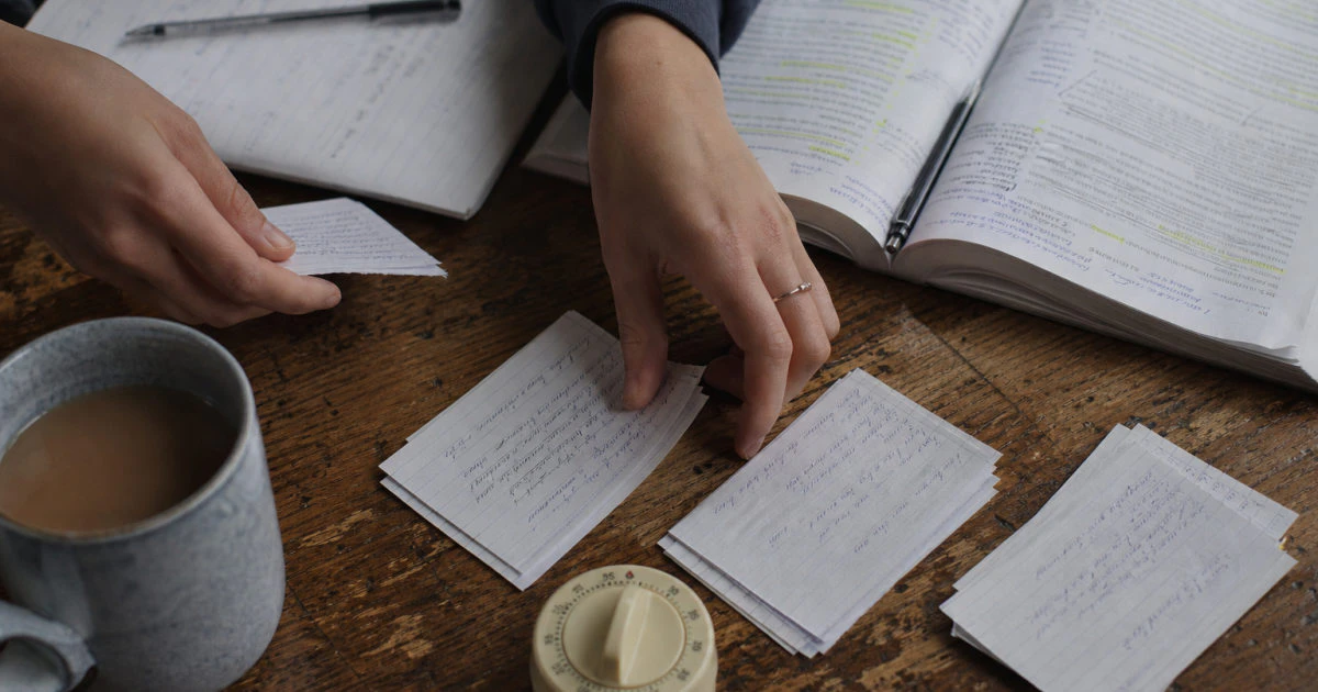 Hands sorting written flashcards beside an annotated textbook, mug, and timer on a kitchen table.