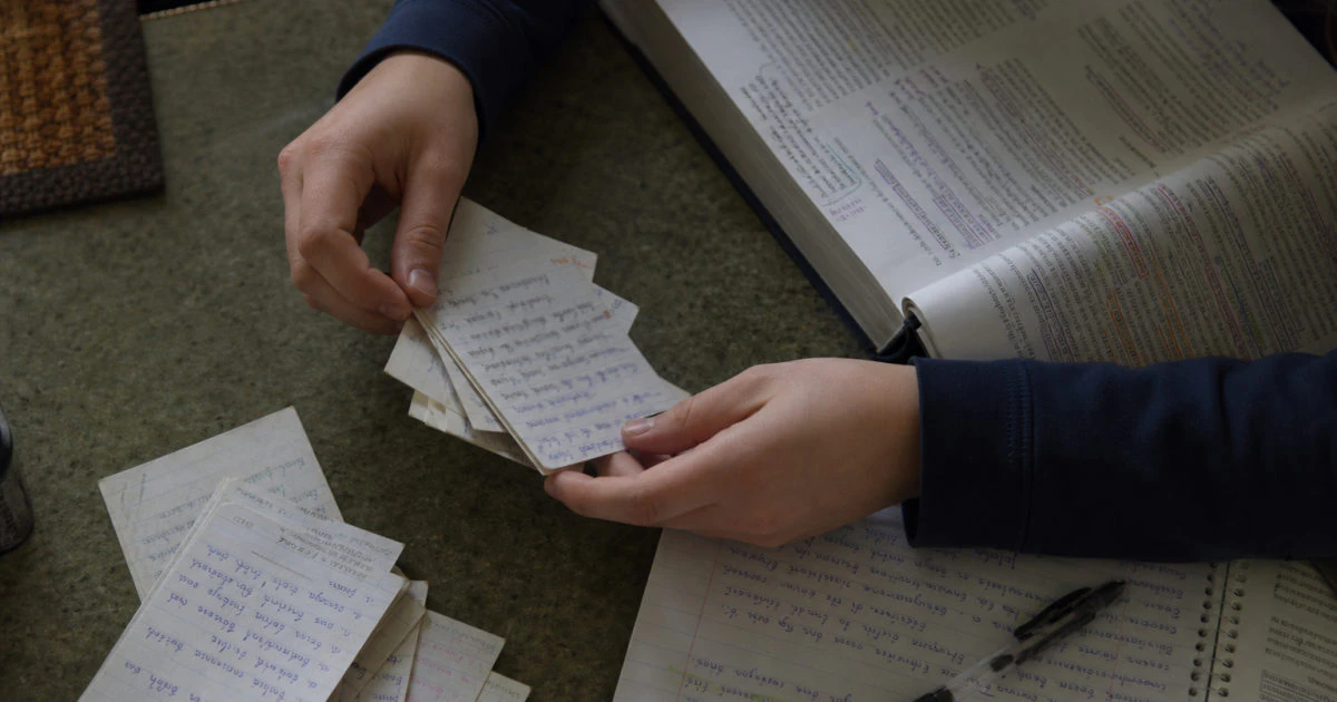 Hands sorting handwritten flashcards beside an annotated biology textbook and notebook on a kitchen table.