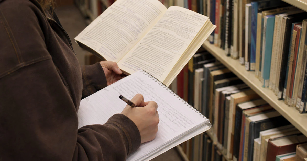 Student in a library aisle writing in a used notebook while holding an annotated textbook open.