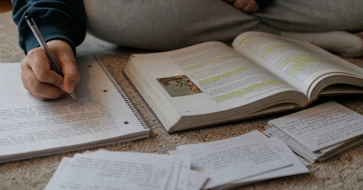 Hands arranging handwritten flashcards beside an open textbook and notebook on a bedroom floor.