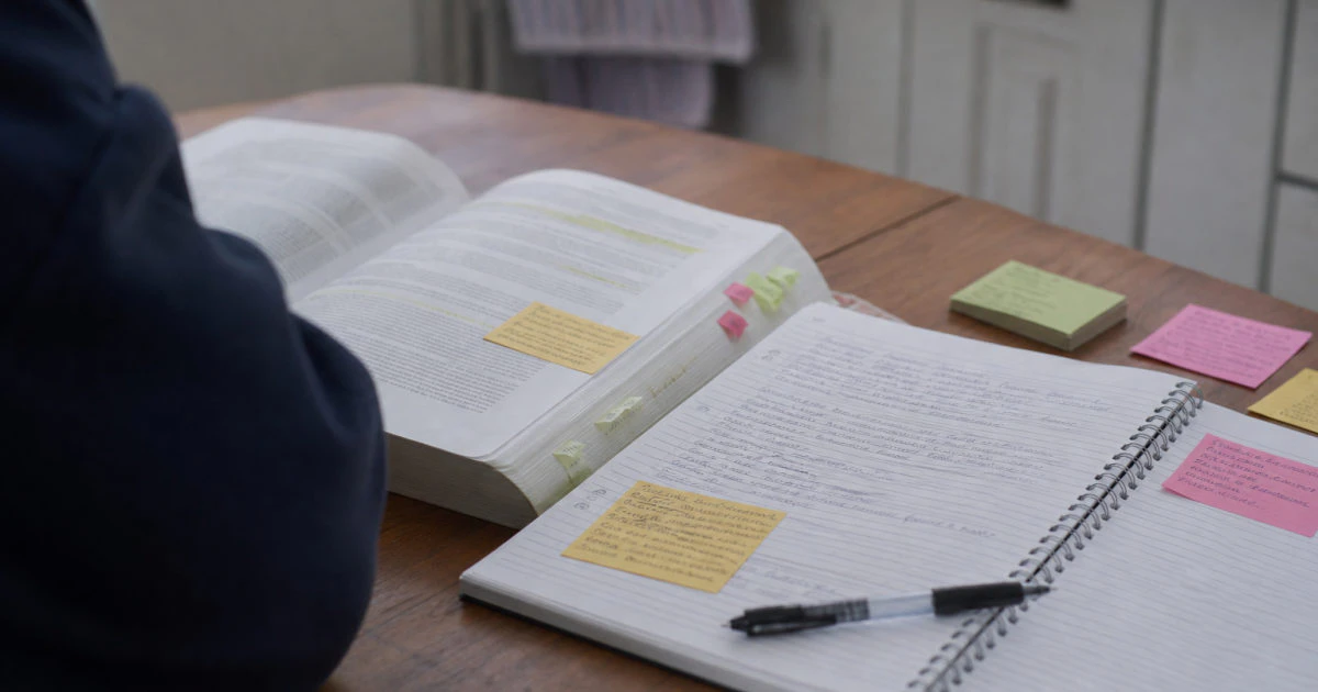 Student at a kitchen table comparing a highlighted textbook with handwritten notes and annotated sticky notes.