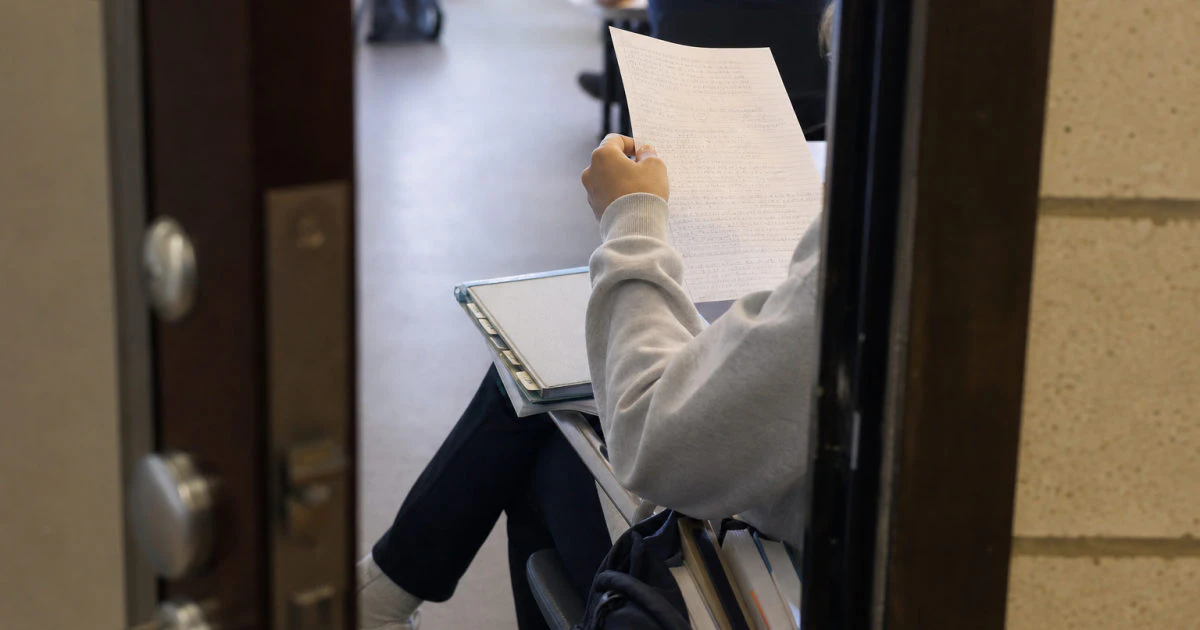 Student seated in a lecture hall corridor reviewing a handwritten summary beside a tabbed textbook and bag of books.