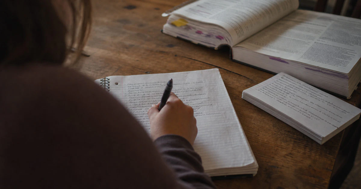 Student writing in a part-filled notebook at a kitchen table with a closed annotated textbook nearby.