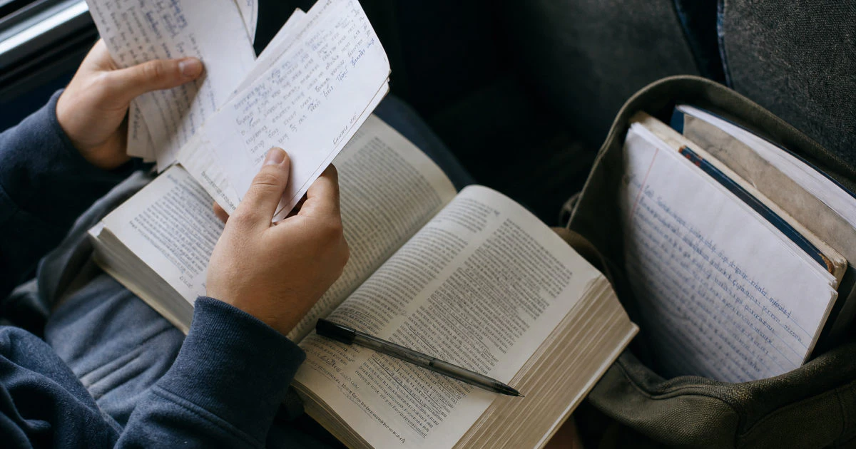 Hands holding written question cards above an open annotated textbook on a bus.