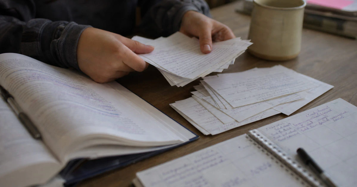 Hands sorting handwritten flashcards beside an annotated textbook and a study calendar on a kitchen table.