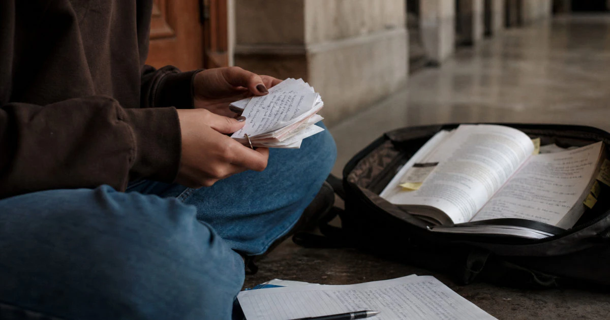 Student seated in a lecture hall corridor reviewing handwritten flashcards beside a bag of annotated books.