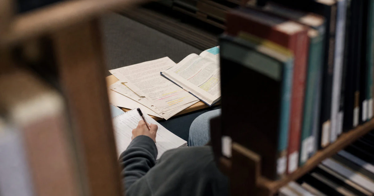 Student sitting between library shelves with printed lecture slides, an open textbook, and a handwritten notebook.
