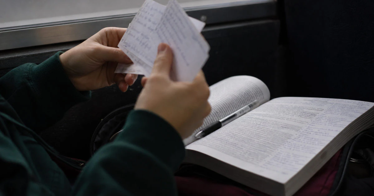 Hands holding handwritten flashcards above an annotated textbook on a bus.