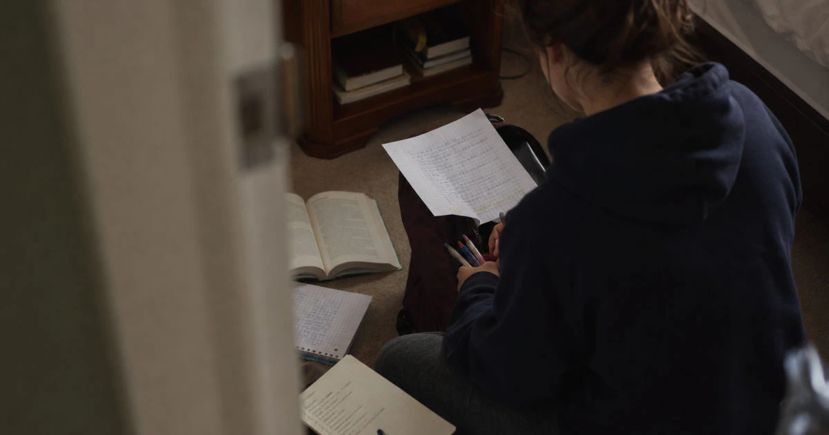 Student packing pens and an annotated review sheet into a bag on a bedroom floor.