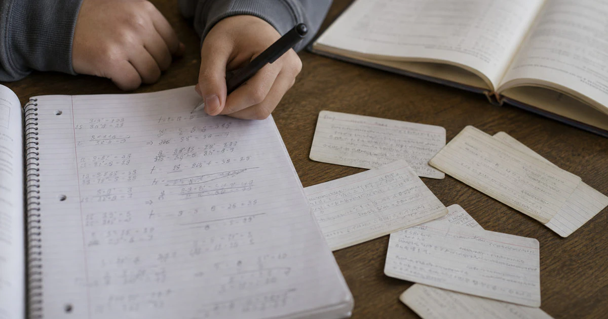 Hands reviewing handwritten formula notes and used study cards beside a completed practice problem on a kitchen table.