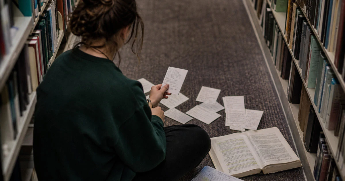 Student viewed from behind sitting in a library aisle with written formula cards, an annotated textbook, and completed problem pages.