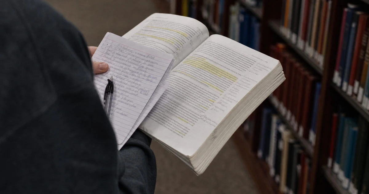 A student in a library aisle holds an annotated textbook and written question cards.
