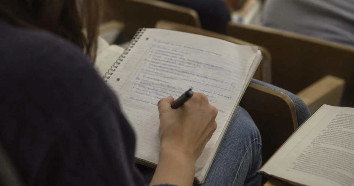 A student writes in a notebook with handwritten notes and question marks in the margin.