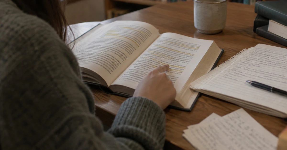 Over-the-shoulder view of a student reading a highlighted textbook beside a notebook with handwritten notes.